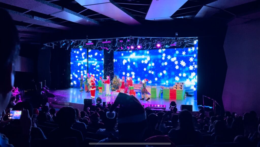 MSCベリッシマのクリスマスクルーズで行われたクリスマスイベントのステージ。サンタクロースとエルフたちが雪の結晶を背景にパフォーマンスし、観客が見守る様子。